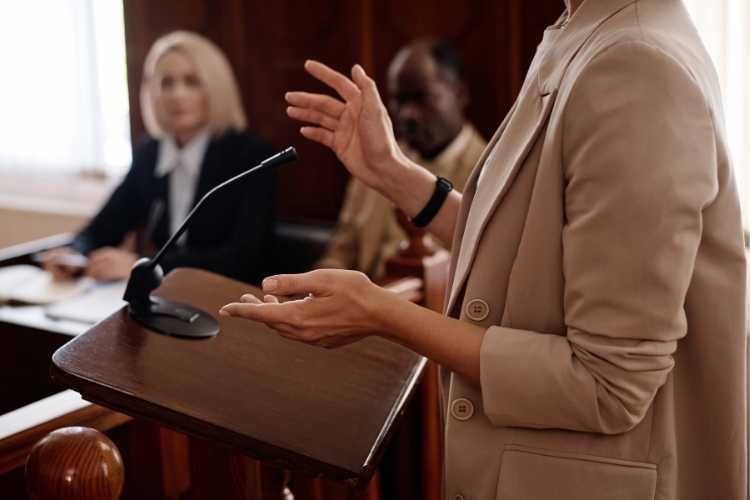 Person testifying at courtroom podium, gesturing by microphone — visual for witness statements service