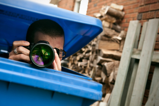 Private investigator hiding in blue bin photographing a subject during a divorce investigation