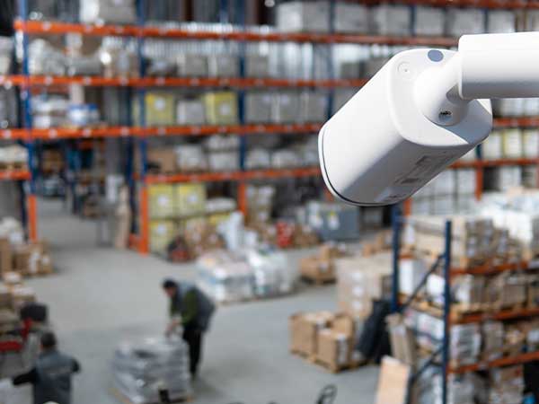 Security camera monitoring warehouse aisles with stacked pallets and workers — business surveillance